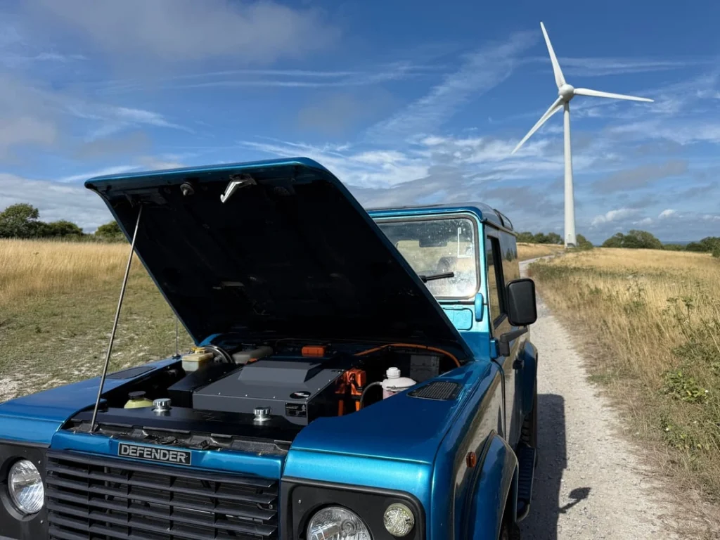 A blue Land Rover Defender 90 with bonnet open, parked near a wind turbine
