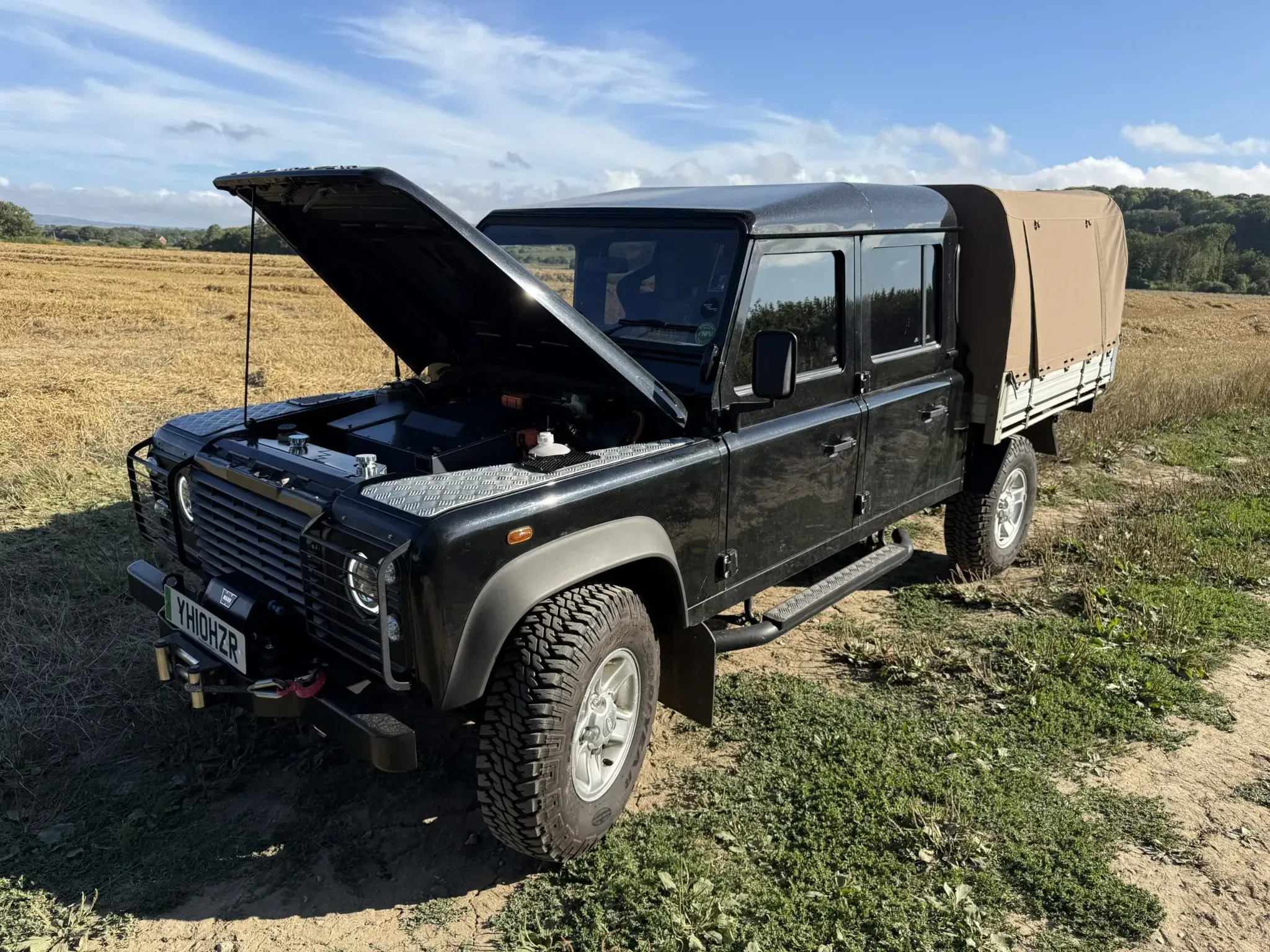 Black Land Rover Defender pickup with open bonnet showing electric conversion in a field.